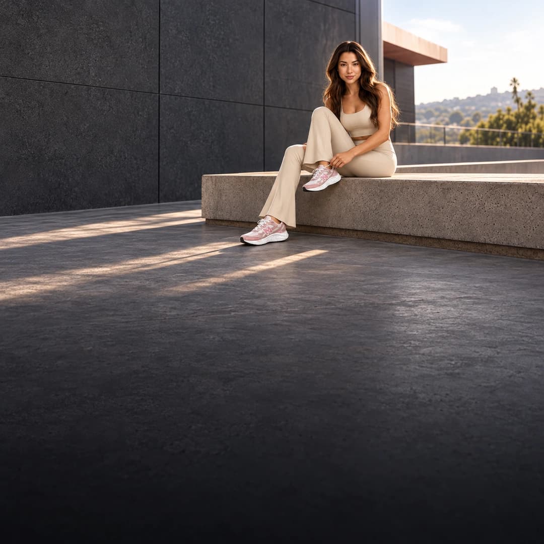 Uma mulher de conjunto bege amarrando o tênis New Balance rosa, sentada num banco de concreto ao sol